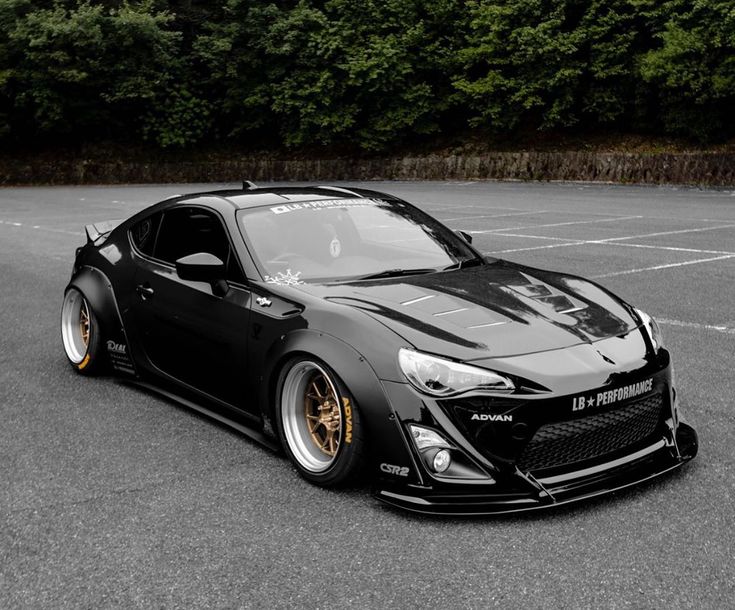 Black sports car toyota 86 with gold rims on a paved surface with greenery in the background
